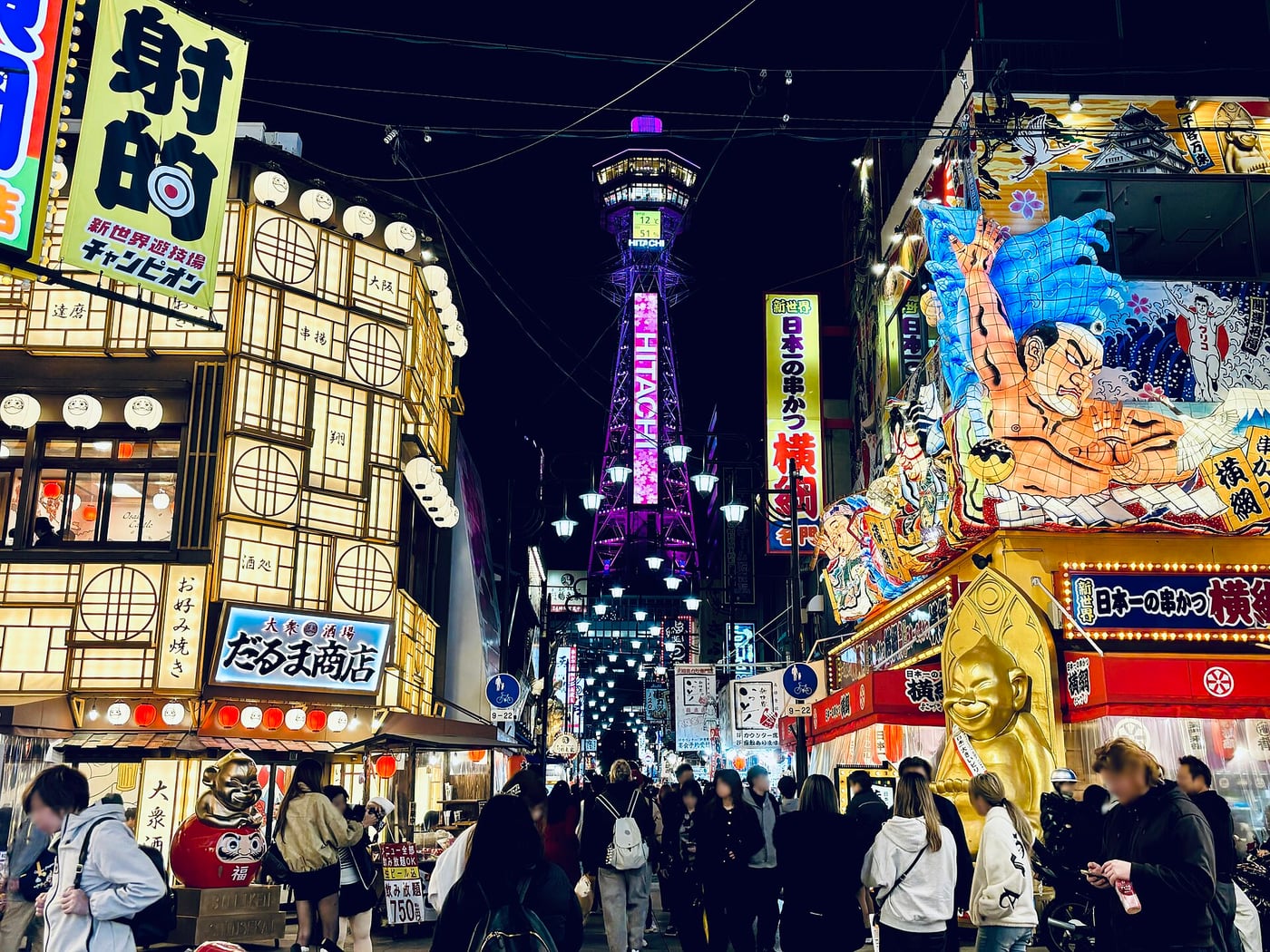 An illuminated Tsutenkaku Tower amid Shinsekai shop signs at night; the itinerary visits during the day